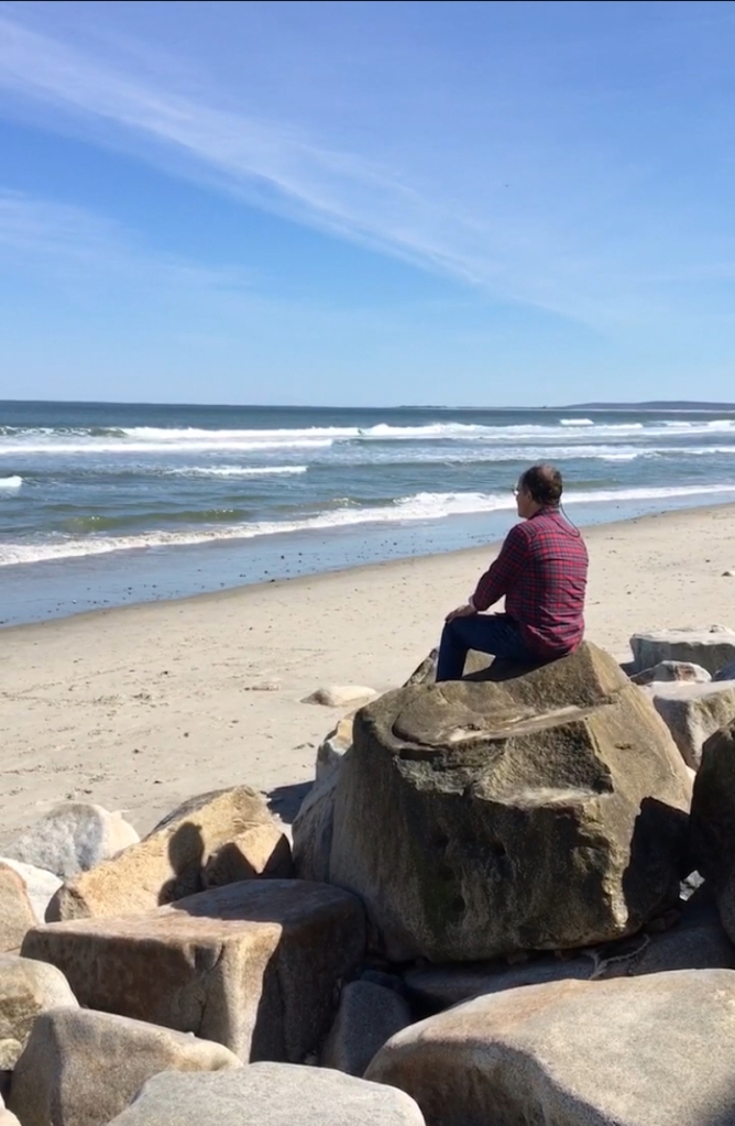 man sitting on beach boulders looking at ocean